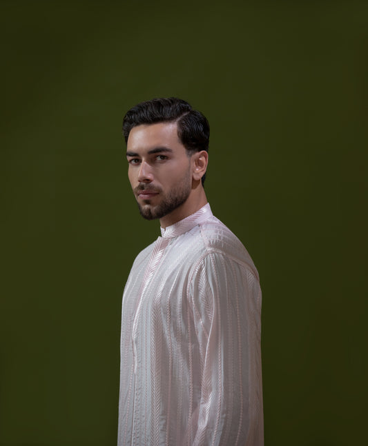 Arsalan Iqbal Blush Pink Chikankari Kurta & Pyjama in Raw Silk Viscose  man wearing a light blush pink full sleeve kurta with all-over white chikankari embroidery on raw silk viscose fabric, mandarin collar with button placket, paired with matching white straight-cut pyjama, styled against an olive green wall with a rustic wooden trunk