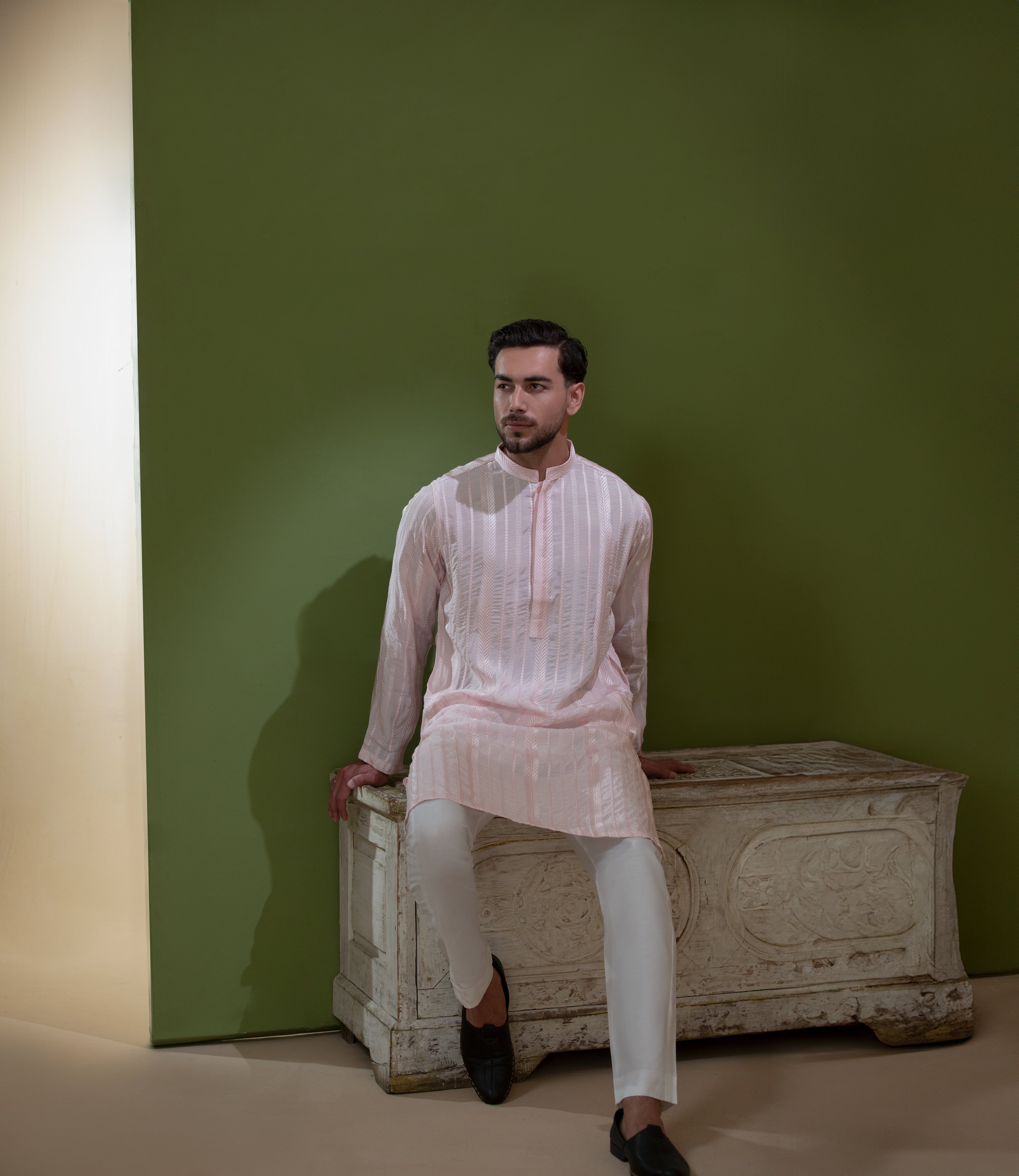 Arsalan Iqbal Blush Pink Chikankari Kurta & Pyjama in Raw Silk Viscose – man wearing a light blush pink full-sleeve kurta with all-over white chikankari embroidery on raw silk viscose fabric, mandarin collar with button placket, paired with matching white straight-cut pyjama, styled against an olive green wall with a rustic wooden trunk