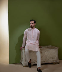 Arsalan Iqbal Blush Pink Chikankari Kurta & Pyjama in Raw Silk Viscose – man wearing a light blush pink full-sleeve kurta with all-over white chikankari embroidery on raw silk viscose fabric, mandarin collar with button placket, paired with matching white straight-cut pyjama, styled against an olive green wall with a rustic wooden trunk