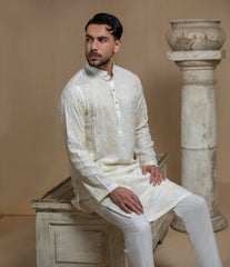 Man wearing Arsalan Iqbal's Cream Kurta Pyjama with allover gold and white floral embroidery, paired with white trousers and a watch, seated against a beige stone backdrop