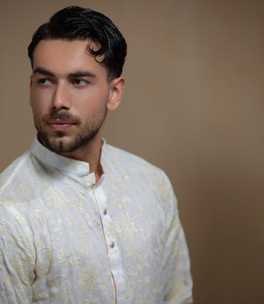 Man wearing Arsalan Iqbal's Cream Kurta Pyjama with allover gold and white floral embroidery, paired with white trousers and a watch, seated against a beige stone backdrop