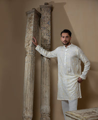 Man wearing Arsalan Iqbal's Cream Kurta Pyjama with allover gold and white floral embroidery, paired with white trousers and a watch, seated against a beige stone backdrop