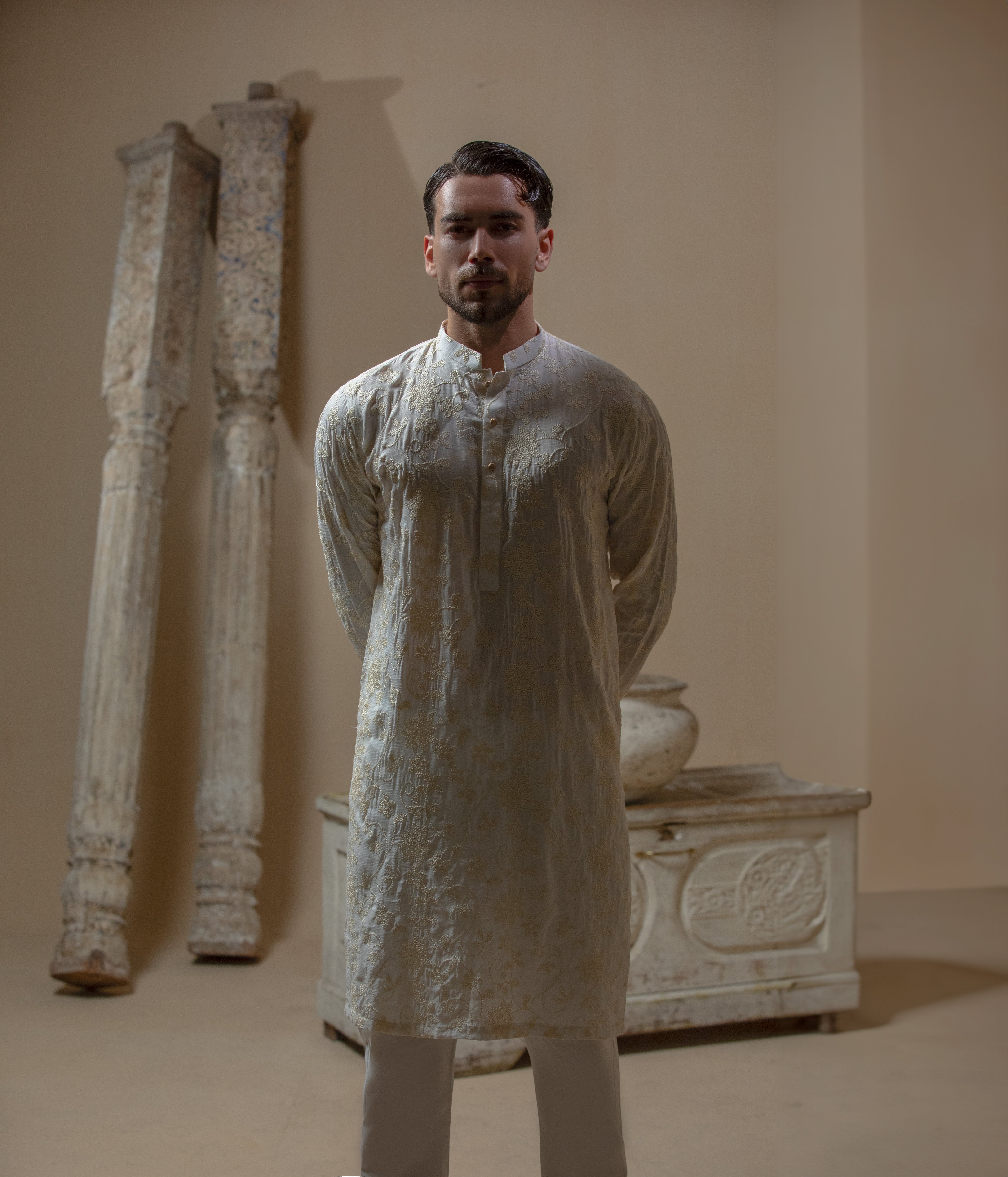 Man wearing Arsalan Iqbal's Cream Kurta Pyjama with allover gold and white floral embroidery, paired with white trousers and a watch, seated against a beige stone backdrop