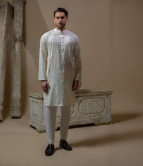 Man wearing Arsalan Iqbal's Cream Kurta Pyjama with allover gold and white floral embroidery, paired with white trousers and a watch, seated against a beige stone backdrop