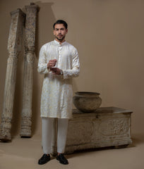 Man wearing Arsalan Iqbal's Cream Kurta Pyjama with allover gold and white floral embroidery, paired with white trousers and a watch, seated against a beige stone backdrop