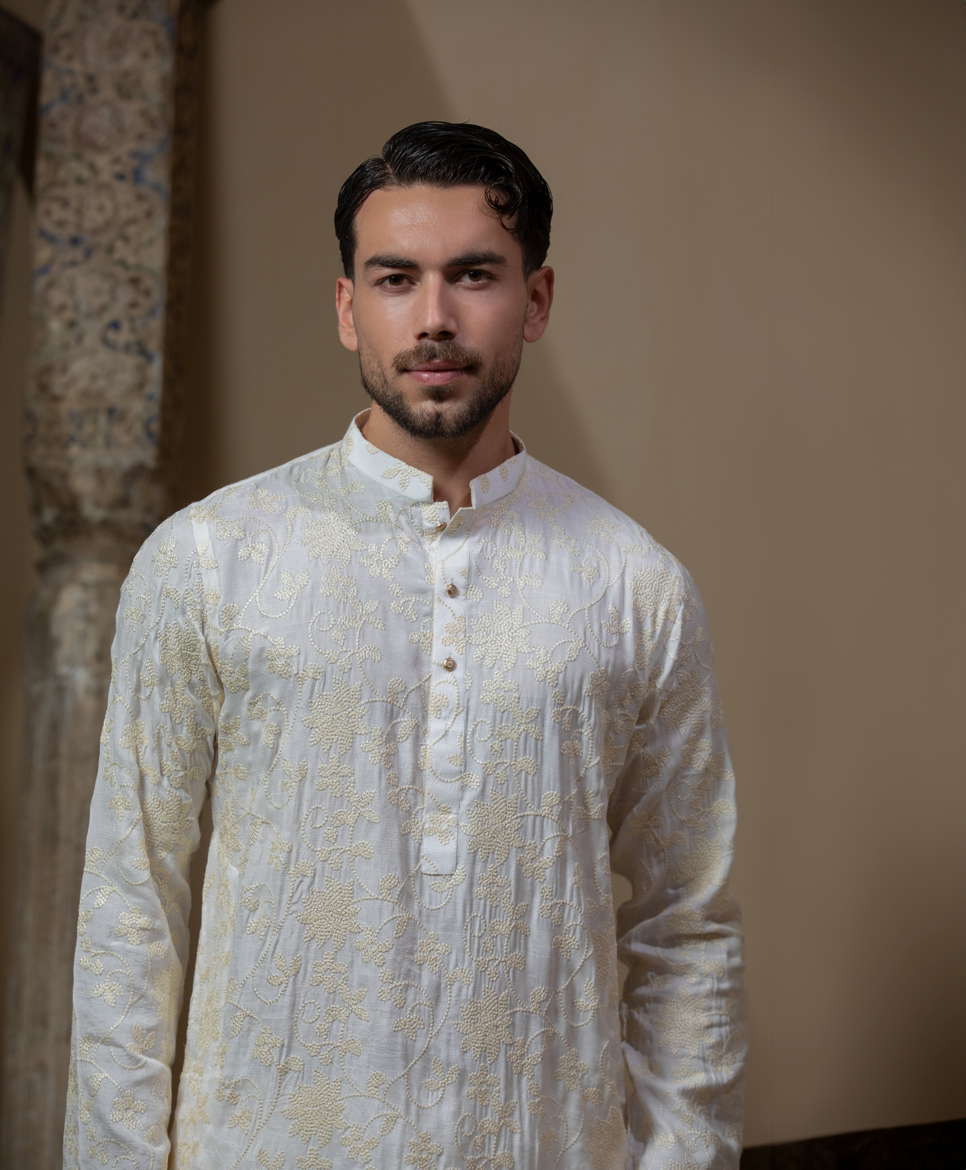 Man wearing Arsalan Iqbal's Cream Kurta Pyjama with allover gold and white floral embroidery, paired with white trousers and a watch, seated against a beige stone backdrop
