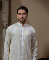 Man wearing Arsalan Iqbal's Cream Kurta Pyjama with allover gold and white floral embroidery, paired with white trousers and a watch, seated against a beige stone backdrop