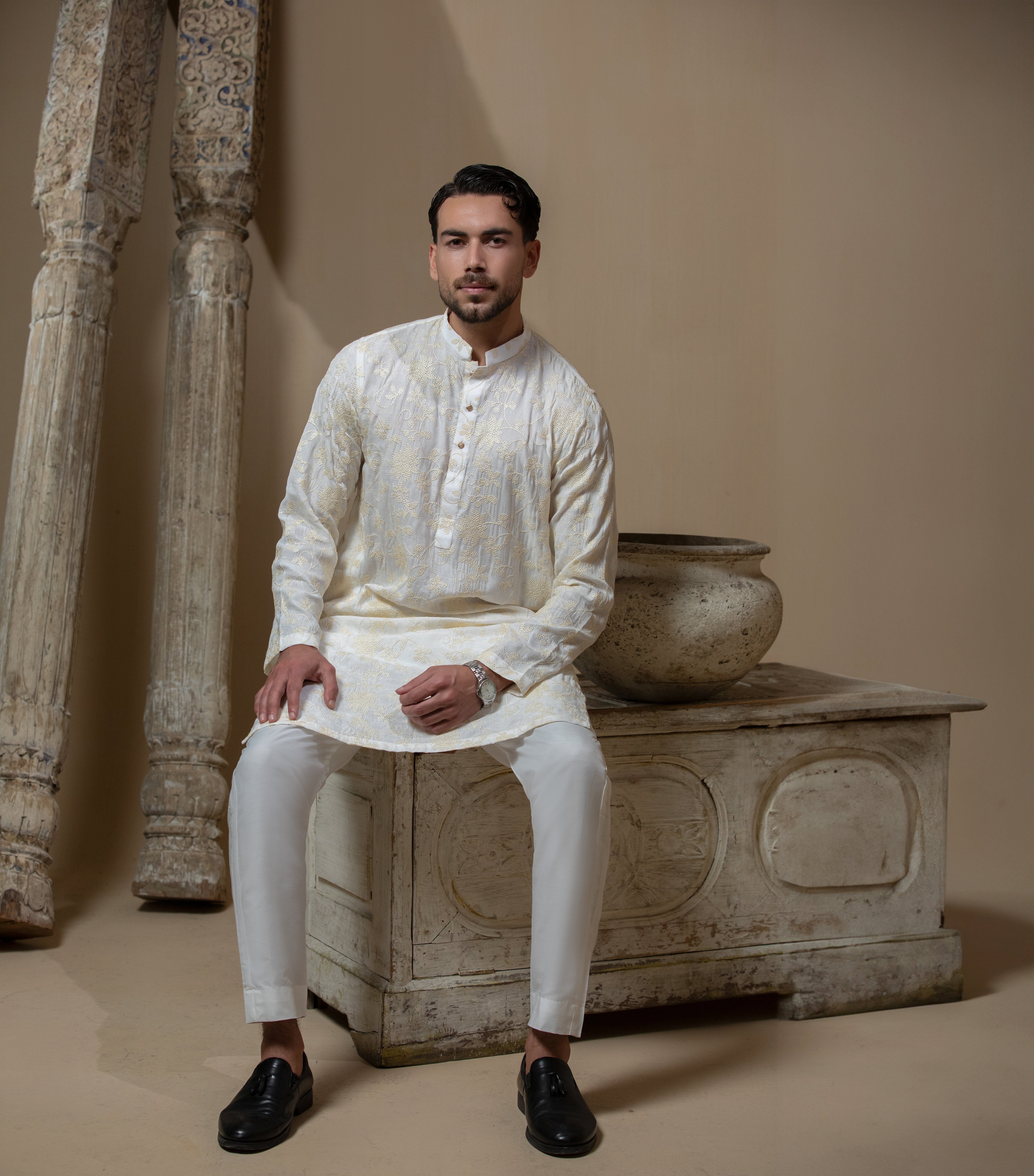 Man wearing Arsalan Iqbal's Cream Kurta Pyjama with allover gold and white floral embroidery, paired with white trousers and a watch, seated against a beige stone backdrop