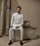 Man wearing Arsalan Iqbal's Cream Kurta Pyjama with allover gold and white floral embroidery, paired with white trousers and a watch, seated against a beige stone backdrop