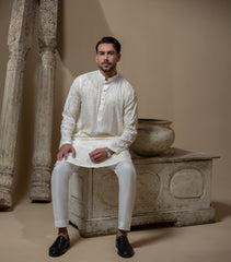 Man wearing Arsalan Iqbal's Cream Kurta Pyjama with allover gold and white floral embroidery, paired with white trousers and a watch, seated against a beige stone backdrop