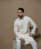Man wearing Arsalan Iqbal's Cream Kurta Pyjama with allover gold and white floral embroidery, paired with white trousers and a watch, seated against a beige stone backdrop