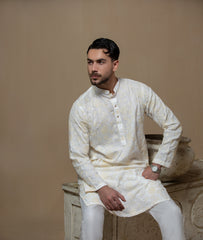 Man wearing Arsalan Iqbal's Cream Kurta Pyjama with allover gold and white floral embroidery, paired with white trousers and a watch, seated against a beige stone backdrop