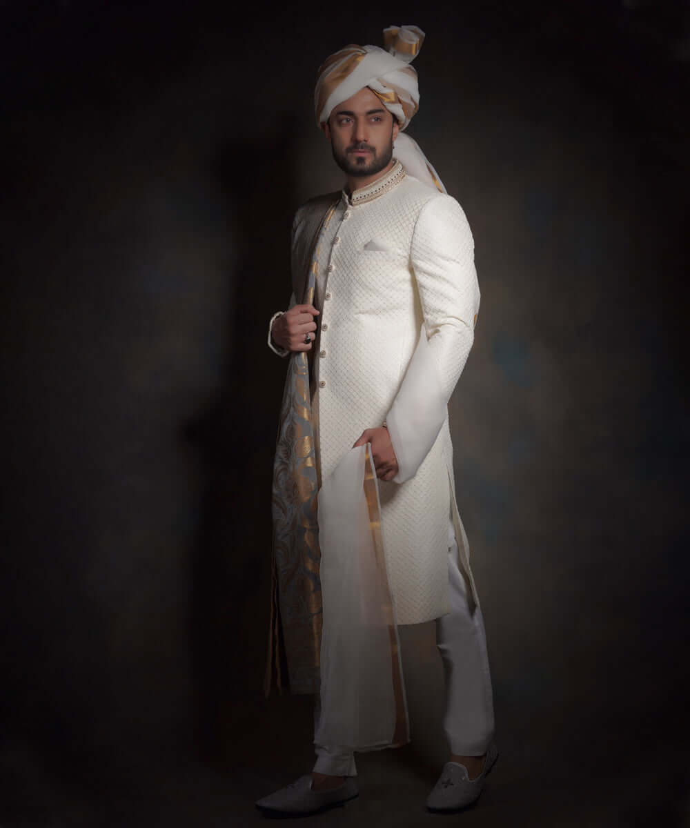 Arsalan Iqbal luxury ivory sherwani with a subtle diamond-textured fabric, featuring an embroidered mandarin collar, styled with a gold and white patterned turban, a matching pocket square, and white formal trousers.
