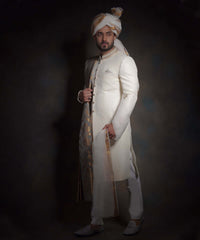 Arsalan Iqbal luxury ivory sherwani with a subtle diamond-textured fabric, featuring an embroidered mandarin collar, styled with a gold and white patterned turban, a matching pocket square, and white formal trousers.