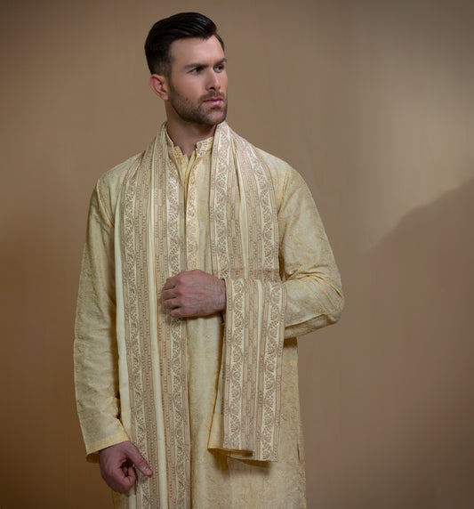 Man wearing Arsalan Iqbal's Light Gold Embroidered Sherwani Coat over a matching kurta pyjama, with a gold-cream stole draped over shoulders and dark formal shoes, posed beside a stone pillar on a beige backdrop