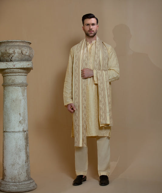 Man wearing Arsalan Iqbal's Light Gold Embroidered Sherwani Coat over a matching kurta pyjama, with a gold-cream stole draped over shoulders and dark formal shoes, posed beside a stone pillar on a beige backdrop