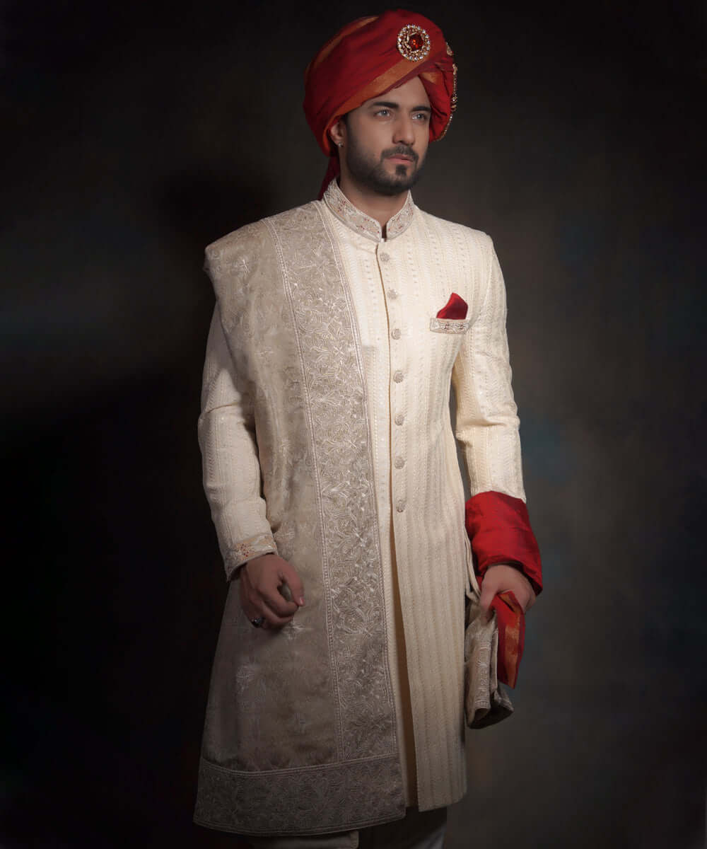 Arsalan Iqbal heritage cream sherwani for grooms with vertical embroidery, paired with a crimson red kulla featuring a jeweled sarpech, an embroidered pocket square, and a draped ivory shawl.