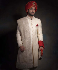 Arsalan Iqbal heritage cream sherwani for grooms with vertical embroidery, paired with a crimson red kulla featuring a jeweled sarpech, an embroidered pocket square, and a draped ivory shawl.