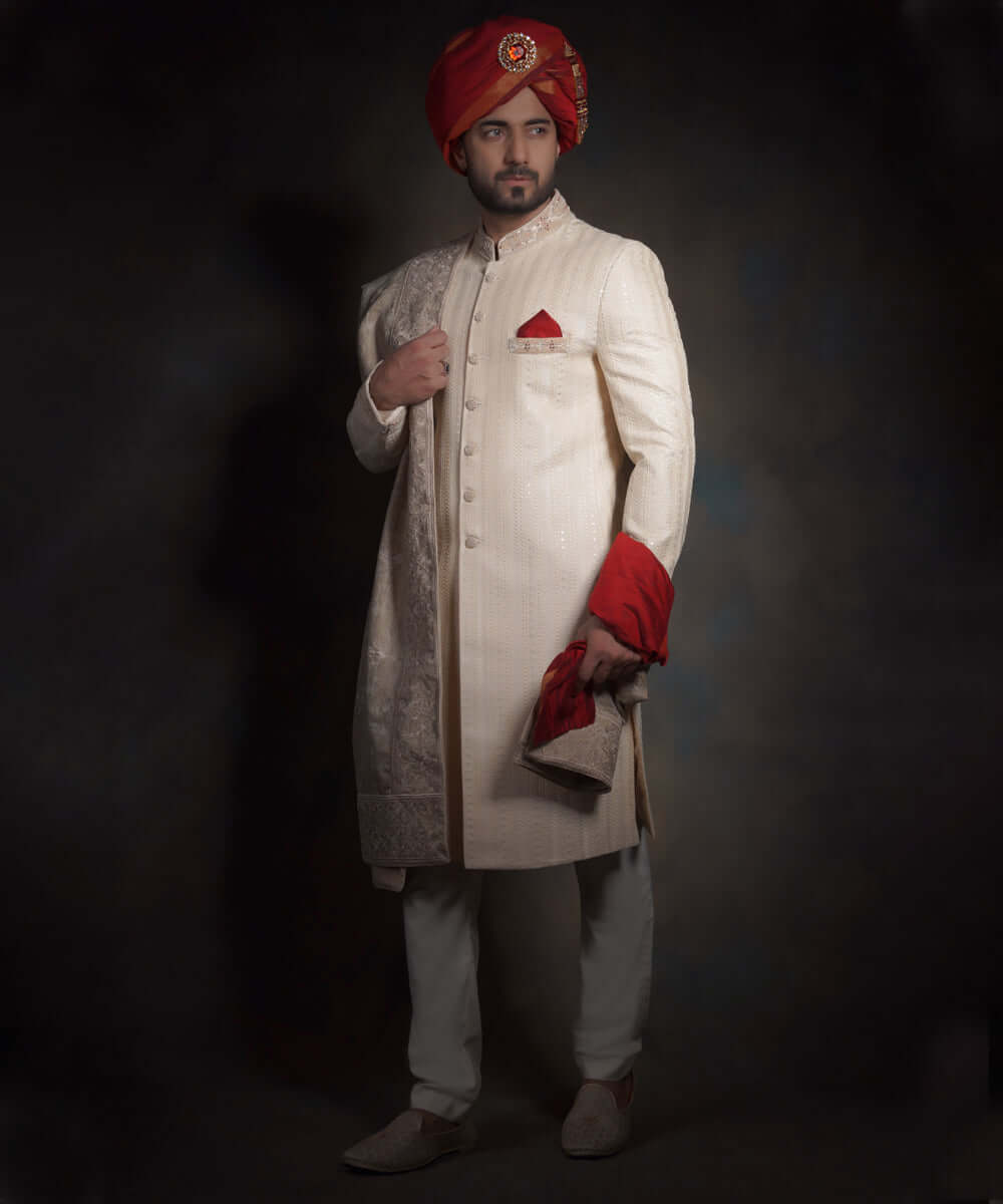 Full-length portrait of Arsalan Iqbal heritage cream sherwani for men, featuring vertical linear embroidery, a crimson red kulla with a jeweled sarpech, an ivory embroidered shawl, and matching traditional mojari shoes.
