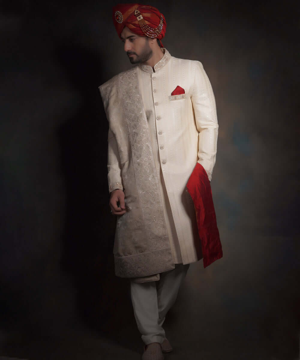 Full-length view of Arsalan Iqbal heritage cream sherwani featuring vertical linear embroidery, styled with a crimson red turban (kulla), a matching red pocket square, ivory trousers, and traditional shoes.
