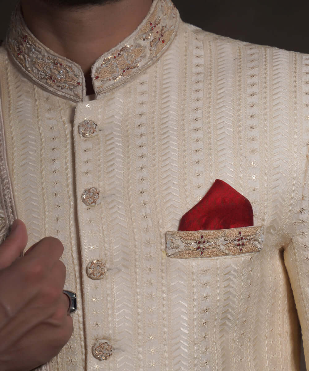 Extreme close-up of Arsalan Iqbal heritage cream sherwani, highlighting vertical linear embroidery, a mandarin collar with gold and red threadwork, fabric-covered buttons, and a red pocket square with an embroidered border.