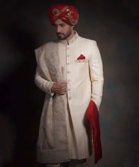 Side-profile portrait of Arsalan Iqbal heritage cream sherwani, featuring a vibrant crimson red kulla with a gold jeweled brooch, a red pocket square, and an intricately embroidered ivory shawl.