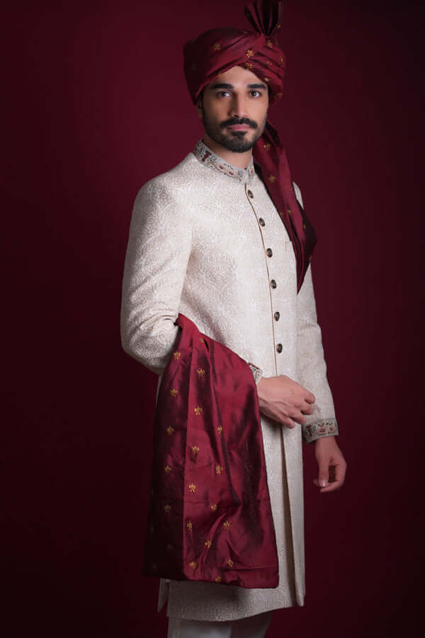 Mid-length portrait of Arsalan Iqbal luxury ivory damask sherwani with a multi-colored paisley embroidered collar, paired with a maroon silk turban and a gold-accented maroon shawl.