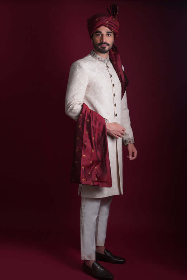 Side profile view of Arsalan Iqbal luxury ivory damask silk sherwani with embroidered collar and cuffs, paired with white trousers, a maroon silk turban, and a gold-accented maroon shawl.