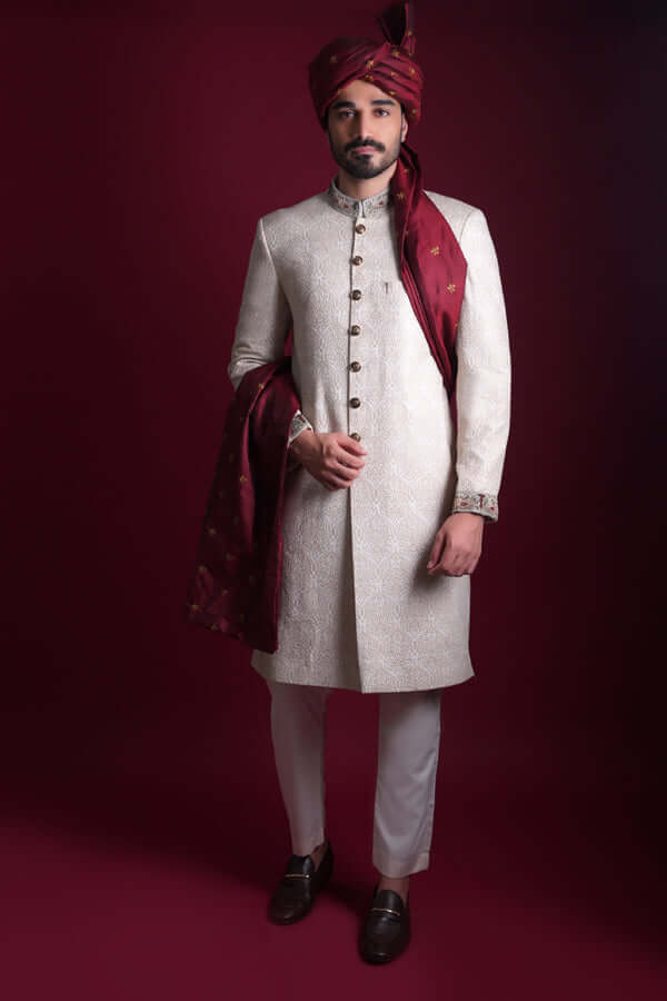 Full-length view of Arsalan Iqbal luxury ivory sherwani with self-on-self damask pattern, featuring embroidered collar and cuffs, styled with a maroon turban and matching silk shawl.