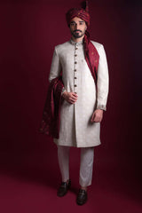 Full-length view of Arsalan Iqbal luxury ivory sherwani with self-on-self damask pattern, featuring embroidered collar and cuffs, styled with a maroon turban and matching silk shawl.