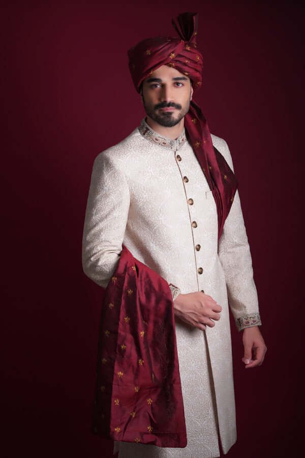 Waist-up portrait of Arsalan Iqbal luxury ivory damask sherwani featuring an embroidered mandarin collar, styled with a grand maroon silk turban and a matching silk shawl with golden embroidery.