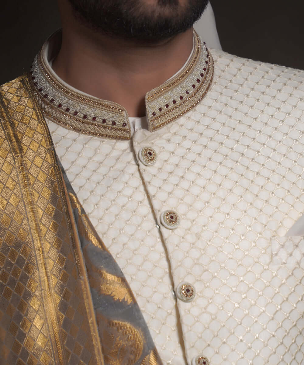 Extreme close-up of Arsalan Iqbal ivory sherwani featuring a raised diamond-textured fabric, an embroidered mandarin collar with gold and maroon accents, and decorative fabric-covered buttons with metallic centers.