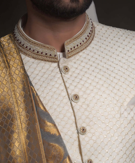 Extreme close-up of Arsalan Iqbal ivory sherwani featuring a raised diamond-textured fabric, an embroidered mandarin collar with gold and maroon accents, and decorative fabric-covered buttons with metallic centers.
