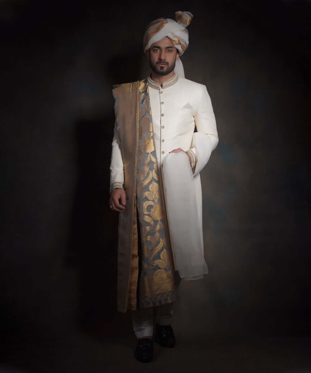 Full-length portrait of Arsalan Iqbal luxury ivory sherwani with diamond-textured fabric, featuring an embroidered mandarin collar, a gold floral silk shawl, matching ivory trousers, and black formal loafers.