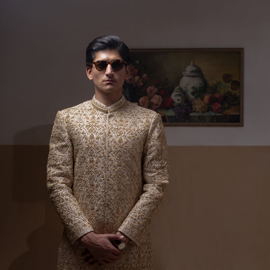 Arsalan Iqbal Ivory & Gold Handcrafted Maharaja Sherwani – man wearing a floor-length ivory sherwani with dense all-over gold hand embroidery featuring intricate floral and botanical motifs, mandarin collar, and a regal structured silhouette, accessorized with sunglasses, styled against a neutral two-tone background with a wooden chair