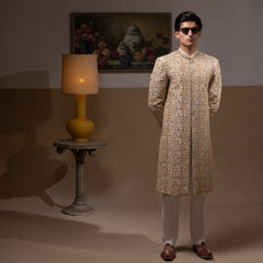 Arsalan Iqbal Ivory & Gold Handcrafted Maharaja Sherwani man wearing a floor-length ivory sherwani with dense all-over gold hand embroidery featuring intricate floral and botanical motifs, mandarin collar, and a regal structured silhouette, accessorized with sunglasses, styled against a neutral two-tone background with a wooden chair