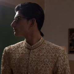 Arsalan Iqbal Ivory & Gold Handcrafted Maharaja Sherwani man wearing a floor-length ivory sherwani with dense all-over gold hand embroidery featuring intricate floral and botanical motifs, mandarin collar, and a regal structured silhouette, accessorized with sunglasses, styled against a neutral two-tone background with a wooden chair