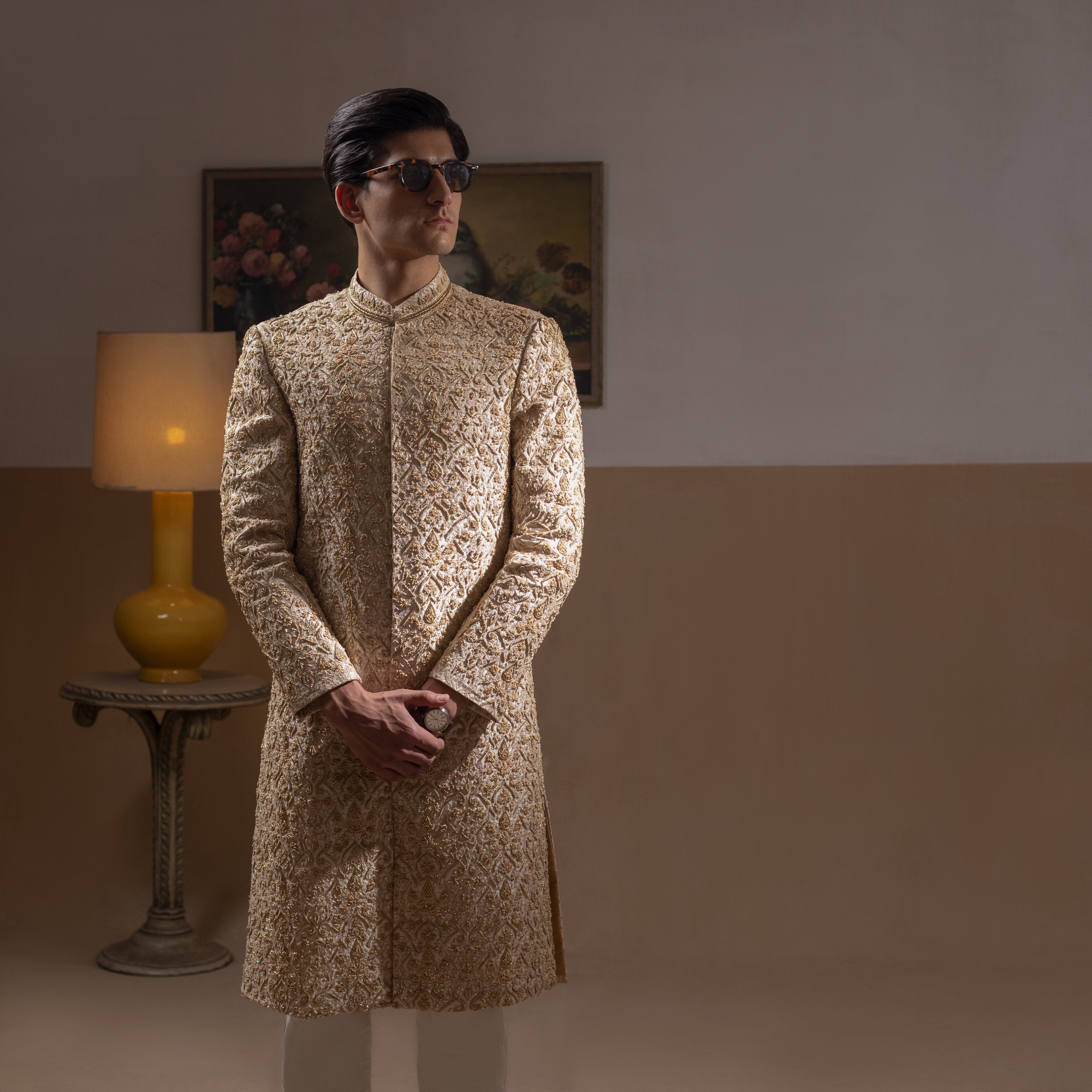 Arsalan Iqbal Ivory & Gold Handcrafted Maharaja Sherwani – man wearing a floor-length ivory sherwani with dense all-over gold hand embroidery featuring intricate floral and botanical motifs, mandarin collar, and a regal structured silhouette, accessorized with sunglasses, styled against a neutral two-tone background with a wooden chair