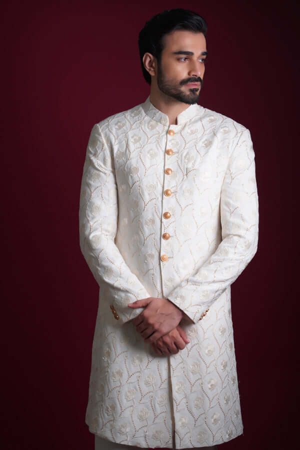 Frontal mid-length portrait of Arsalan Iqbal luxury ivory sherwani with all-over white floral embroidery, featuring gold buttons and a mandarin collar, with model looking to the side.
