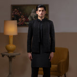 Arsalan Iqbal Jet Black Hand Embroidered Prince Jacket with Kurta Pyjama man wearing a fully embellished jet black prince jacket featuring dense hand embroidery with sequin and thread detailing, paired with a matching black kurta pyjama, mandarin collar, and a sleek formal silhouette, styled in a warm indoor setting