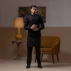 Arsalan Iqbal Jet Black Hand Embroidered Prince Jacket with Kurta Pyjama  man wearing a fully embellished jet black prince jacket featuring dense hand embroidery with sequin and thread detailing, paired with a matching black kurta pyjama, mandarin collar, and a sleek formal silhouette, styled in a warm indoor setting