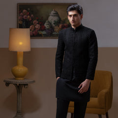 Arsalan Iqbal Jet Black Hand Embroidered Prince Jacket with Kurta Pyjama – man wearing a fully embellished jet black prince jacket featuring dense hand embroidery with sequin and thread detailing, paired with a matching black kurta pyjama, mandarin collar, and a sleek formal silhouette, styled in a warm indoor setting