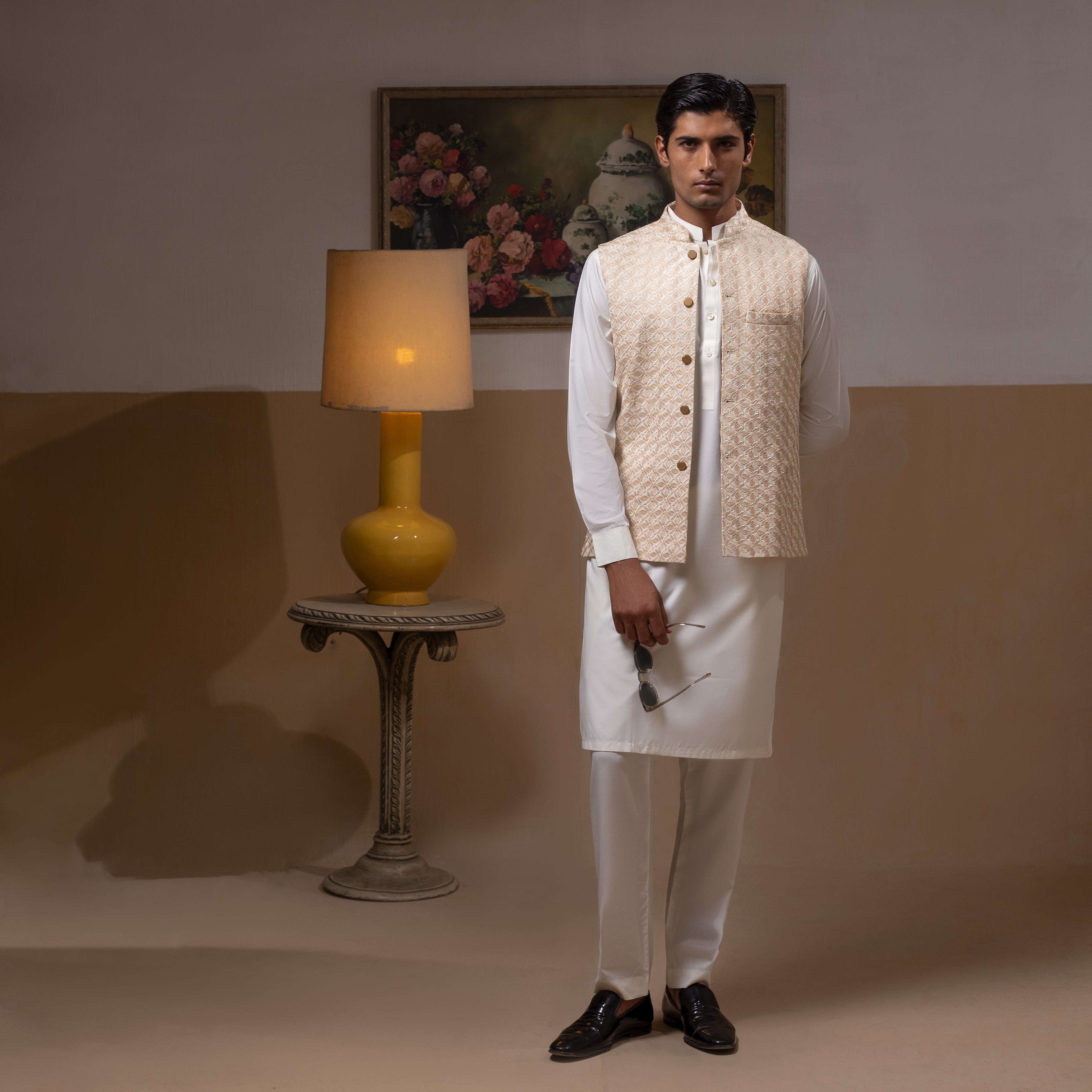 Arsalan Iqbal Light Gold Embroidered Waistcoat with IronEZ Kurta & Pyjama Set – man wearing a light gold heavily embroidered sleeveless waistcoat with gold button closure over a crisp white full-sleeve kurta, paired with matching white straight-cut pyjama, accessorized with sunglasses, styled in a warm classic indoor setting