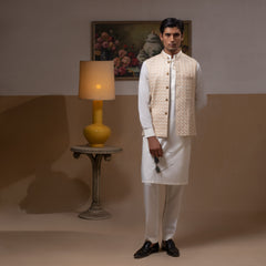 Arsalan Iqbal Light Gold Embroidered Waistcoat with IronEZ Kurta & Pyjama Set – man wearing a light gold heavily embroidered sleeveless waistcoat with gold button closure over a crisp white full-sleeve kurta, paired with matching white straight-cut pyjama, accessorized with sunglasses, styled in a warm classic indoor setting