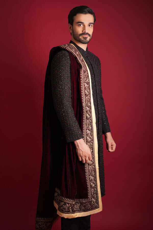 Side profile of Arsalan Iqbal black sherwani with red beadwork, paired with a draped maroon velvet shawl featuring a wide gold tilla border, styled against a crimson background.