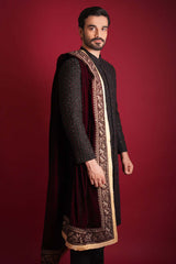 Side profile of Arsalan Iqbal black sherwani with red beadwork, paired with a draped maroon velvet shawl featuring a wide gold tilla border, styled against a crimson background.