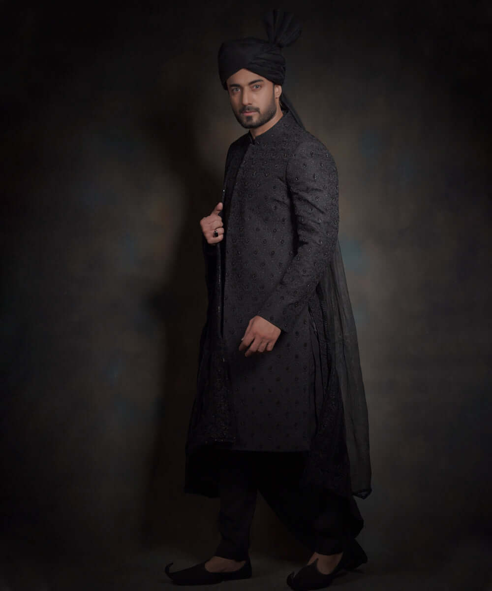 Side profile view of Arsalan Iqbal midnight black luxury sherwani featuring spaced-out tonal motifs, styled with a high-pleated black turban, matching trousers, and traditional khussa shoes.
