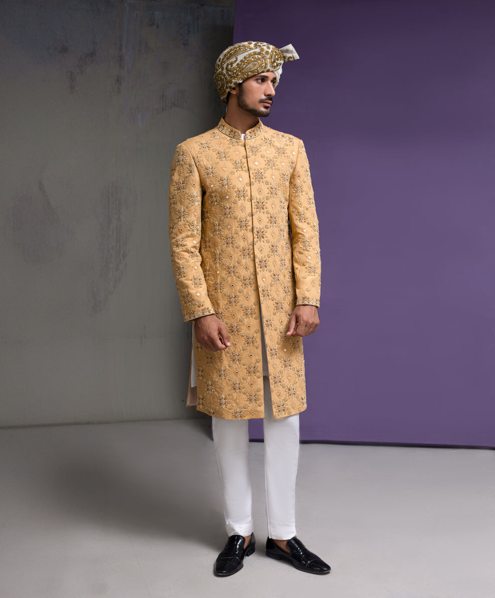 Arsalan Iqbal mustard gold groom sherwani featuring intricate all-over floral embroidery and mirror-work, paired with white trousers and a traditional patterned turban in a full-length studio pose.