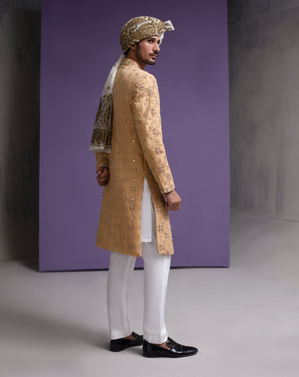 Side profile view of an Arsalan Iqbal mustard gold luxury sherwani for men, featuring intricate floral embroidery, mirror-work accents, a patterned white and gold turban, white trousers, and black loafers.