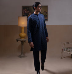 Man wearing Arsalan Iqbal's Navy Blue Allover Sequin Embroidered Kurta with dark navy trousers, black loafers and sunglasses, posed against a beige backdrop with yellow lamp and floral painting
